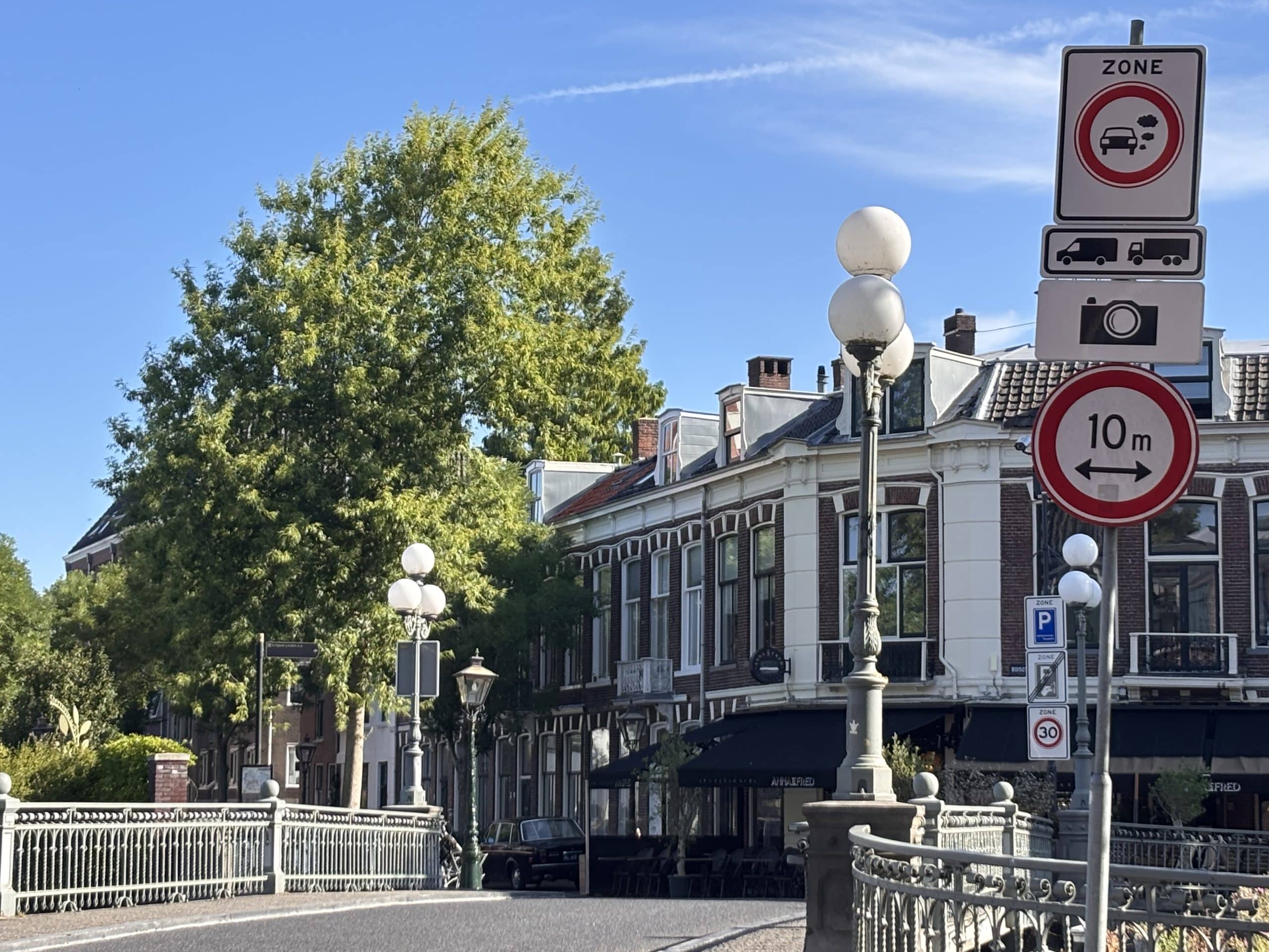 Afbeelding van een Leidse straat met brug waarnaast een paal met het nieuwe verkeersbord staat. Het bovenste bord is wit, met de tekst ‘ZONE’ en een rode cirkel met daarin een auto die uitlaatgassen uitstoot. Dit geeft aan dat een voertuig een gebied binnenrijdt waar toegang beperkt is voor voertuigen met uitstoot. Daaronder hangt een bord waarop staat welke voertuigen geen toegang hebben tot deze zone. Dat zijn in Leiden een bestel- en vrachtauto.